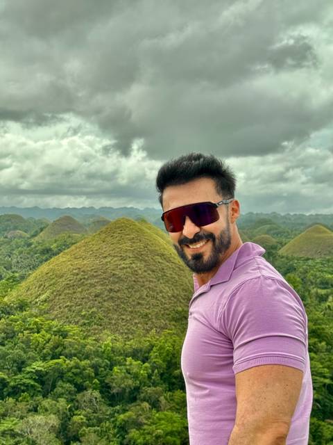       Man wearing sunglasses smiles with Bohol’s Chocolate Hills rolling across a cloudy tropical landscape behind him.
  