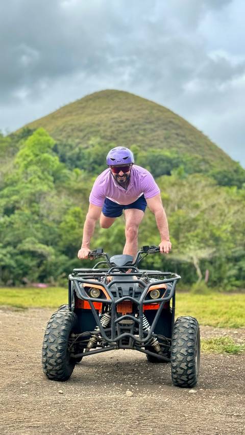       Man wearing helmet performs a balancing stunt on an off-road vehicle platform with jungle backdrop.
  