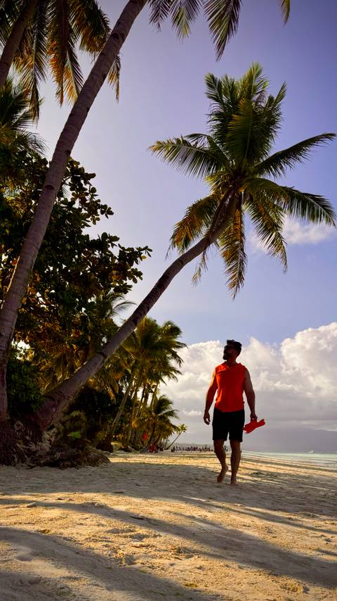       Silhouetted man looks up at tall leaning palm trees against a bright sky with fluffy clouds.
  