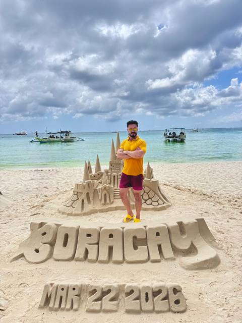       Man in bright outfit poses proudly beside an elaborate sandcastle on a sunny tropical beach with boats offshore.
  