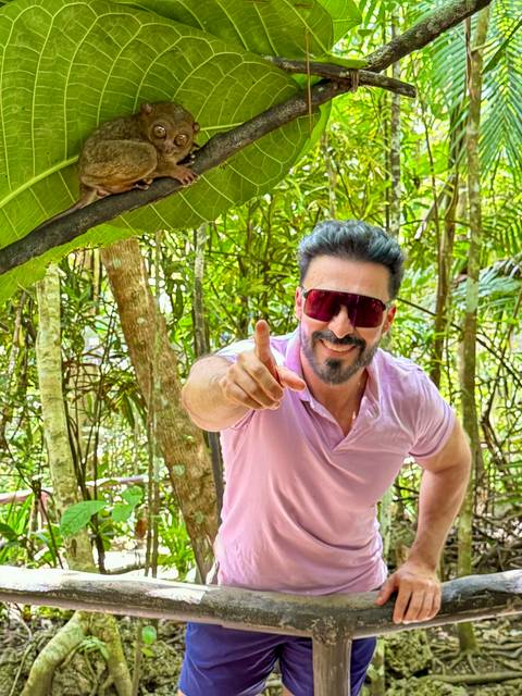       Man wearing reflective sunglasses playfully points toward the camera in a dense tropical garden.
  