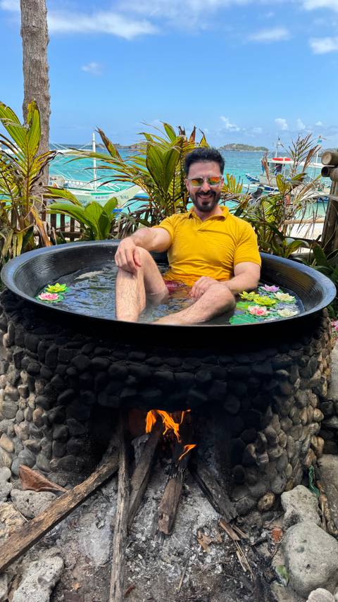       Traveler lounges in an outdoor stone tub filled with water and floating lotus flowers in a tropical setting.
  