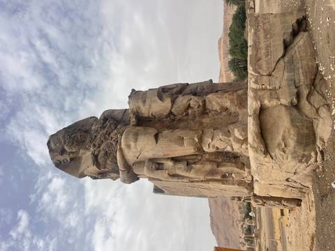       Massive seated Colossus statue carved from sandstone stands under partly cloudy skies in Luxor.
  