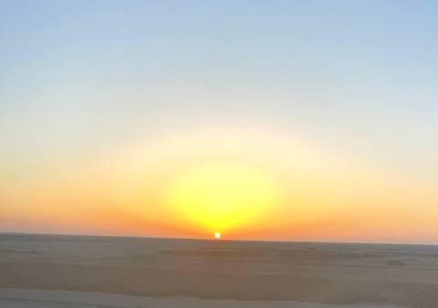       Sun dips below a vast desert horizon creating a glowing orange halo in the sky.
  