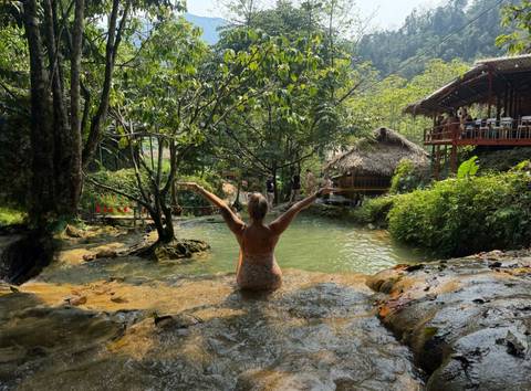       Traveler sits waist-deep in a natural spring pool surrounded by lush vegetation, arms outstretched in joy.
  