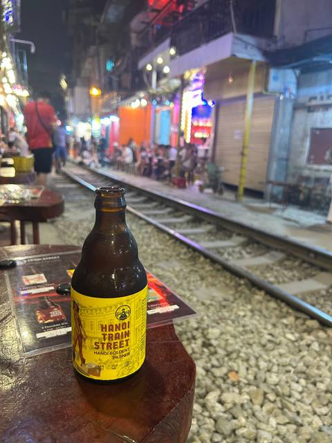      Beer bottle in focus on Hanoi’s famous Train Street with blurred crowds and railway behind.
  
