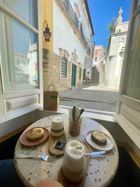       Cozy café table with pastry and coffee by an open window overlooking a sunlit stone alley and staircase.
  