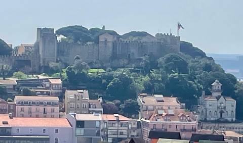       Distant view of São Jorge Castle crowning a wooded hill above Lisbon rooftops.
  