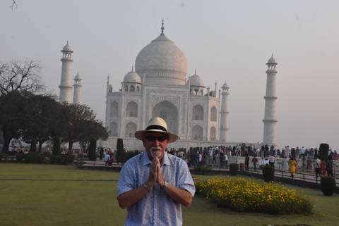       Smiling traveller posing with hands in prayer in front of the hazy white marble Taj Mahal.
  