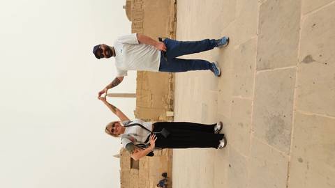       Two travellers perform a playful perspective pose touching the tip of a distant stone obelisk in an ancient temple courtyard.
  