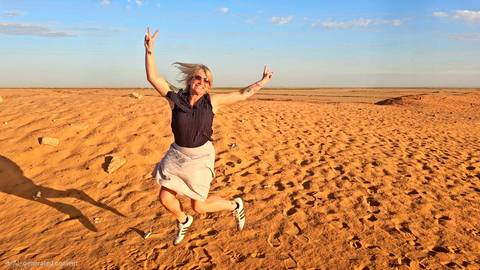       A joyful woman jumps mid-air on a vast sandy desert plain under a bright blue sky.
  