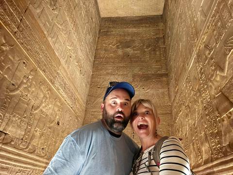       Wide-angle selfie of two travellers making exaggerated faces inside a narrow hieroglyph-covered chamber.
  