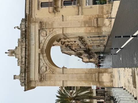       A tall limestone triumphal arch with eagle statue towers over a wet street in a historic town.
  
