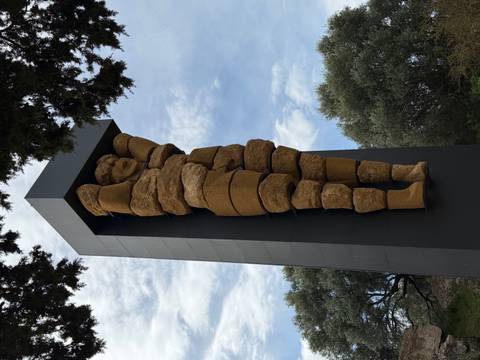       Modern stacked-stone sculpture of an atlas figure stands against a partly cloudy sky amid olive trees.
  