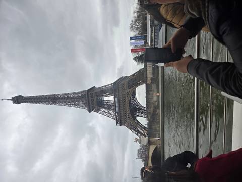       Daytime view of the Eiffel Tower rising over the Seine River captured from a boat, with a visitor taking a photo.
  