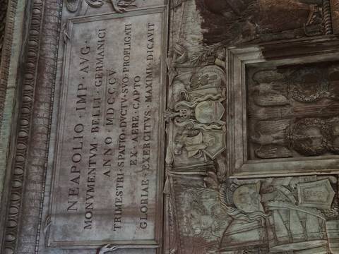       Close-up of Latin inscriptions and sculpted reliefs on a historic triumphal arch.
  