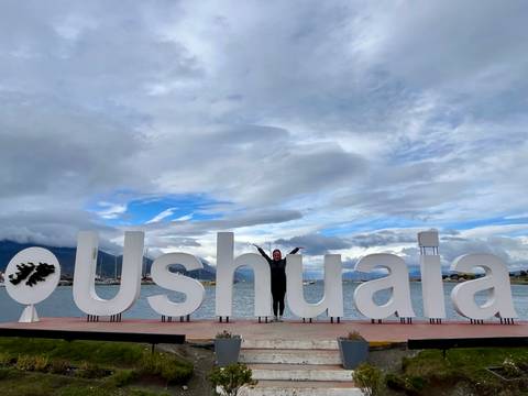       A traveller poses with outstretched arms in front of the oversized white "Ushuaia" sign by the waterfront with mountains and clouds behind.
  