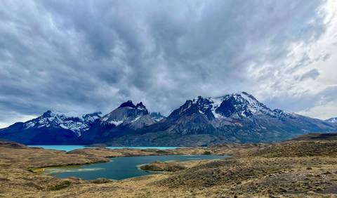       Dramatic snow-capped mountains tower over turquoise lakes and rolling steppe under moody clouds in Patagonia.
  