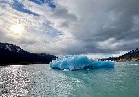       An intense blue iceberg floats serenely in emerald water beneath a bright sun peeking through clouds and snowy peaks.
  