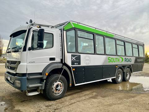       A rugged expedition bus with green and white livery is parked on a gravel lot under a grey sky.
  