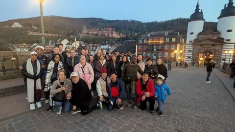       Large tour group gathered on Heidelberg’s Old Bridge with the illuminated castle and town backdrop at dusk.
  