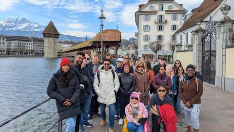       A smiling tour group stands beside Lucerne’s Chapel Bridge and Water Tower with snowy Alps behind.
  