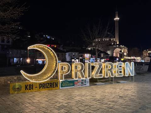       Illuminated crescent-moon and PRIZREN sign shining in a central square at night with a mosque in the background.
  