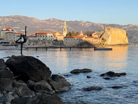       Coastal fortress town with dancer statue on a rock in the foreground and mountains beyond the bay.
  