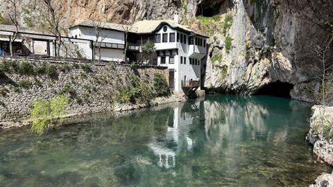       White dervish house nestled beside a turquoise spring emerging from a rocky cave mouth.
  