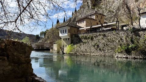       Historic riverside complex of stone buildings and cypress trees mirrored in crystal clear water under a bright sky.
  