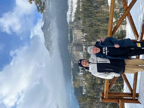       Two travellers smiling with snow-covered Banff Springs Hotel and forested mountains behind.
  