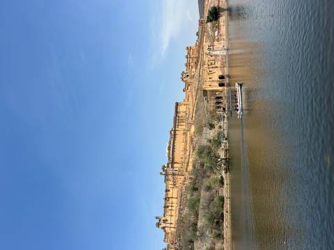       Amber Fort sits majestically above a tranquil lake while a small tourist boat crosses the water under clear blue skies.
  
