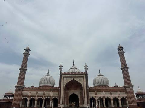       Grand facade of Jama Masjid with three white domes and towering minarets under a cloudy sky.
  