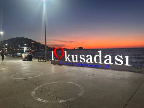       Sunset colours behind the illuminated ‘I ♥ Kusadasi’ seafront sign beside the Aegean Sea.
  
