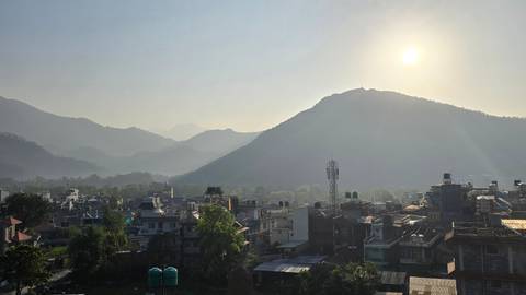       Hazy morning sun over Pokhara’s rooftops with rolling green hills and distant peaks.
  
