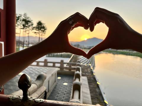       Hands forming a heart shape framing the setting sun over a tranquil river and distant mountain.
  