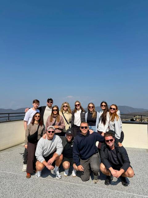       Large group of travelers posing on a terrace with a faint snow-capped Mount Fuji in the background under a clear sky.
  