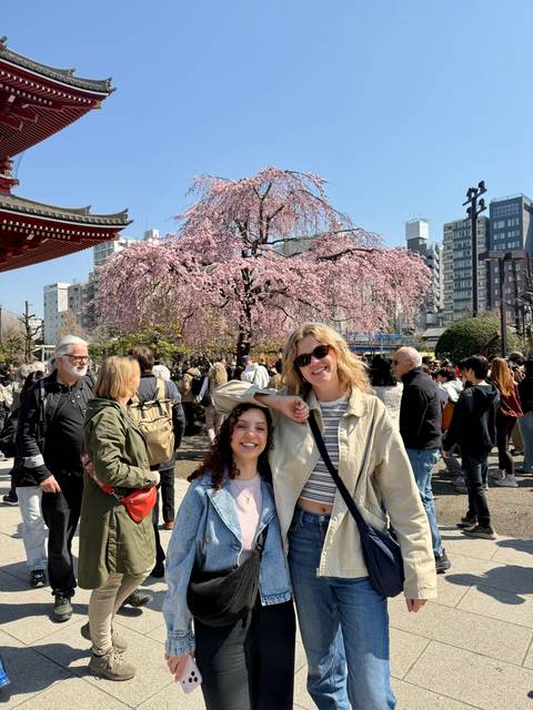       Two women smiling under a cherry blossom tree amid crowds at a city temple fair.
  