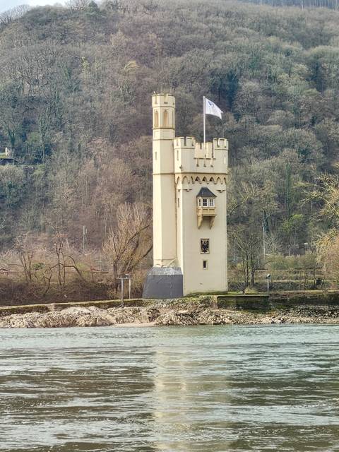       Tall cream‐coloured medieval watchtower rising beside a river with wooded hills in the background.
  