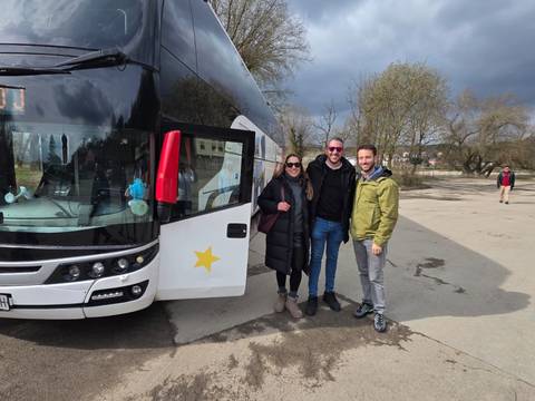       Tour passengers smiling beside a white coach bus with a star logo on a cloudy day.
  