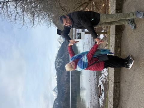       Smiling couple forming a heart shape with their arms beside a calm alpine lake backed by snowy mountains.
  