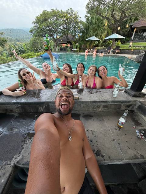       A group of friends cheers with drinks while relaxing in a poolside bar, snapping a fun selfie.
  