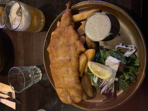       A classic British pub meal of golden fish and chips with tartar sauce and salad sits on a dark wooden table.
  