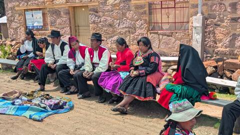      Local men and women in traditional Andean dress sitting on a bench outside a stone building.
  