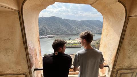       Two young men lean on a railing inside a stone window arch overlooking a lake and green hills.
  