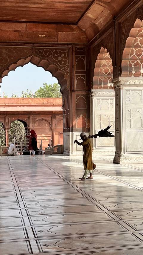       Inside courtyard of historic mosque where a worker sweeps the marble floor with a large broom.
  