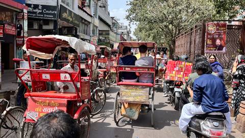       Congested market lane filled with red cycle-rickshaws, scooters and shoppers under bright sun.
  