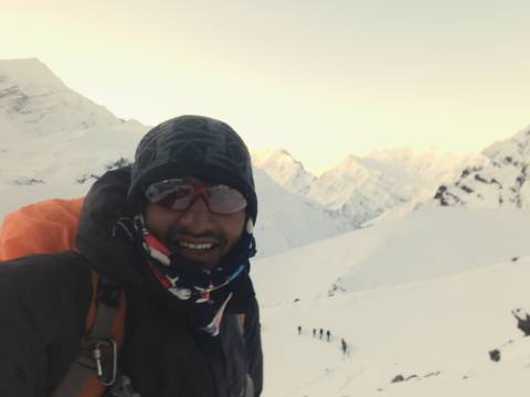       Selfie of bundled trekker smiling with snowy ridges glowing in soft dawn light behind.
  