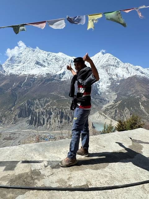       Trekker points triumphantly at the towering snow mountains from a rocky lookout.
  
