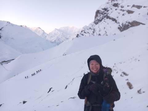       Smiling trekker in winter gear on snowy trail with line of hikers behind in soft dawn light.
  